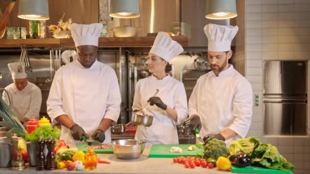 三个穿着白色制服烹饪的专业迷人厨师在厨房边聊天边微笑的画像 白人和非洲裔美国人的主厨们在切蔬菜 烹调概念 — 图库视频影像