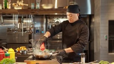 Siyah üniformalı yakışıklı Asyalı aşçının portresi ve tavayla lezzetli yemekler pişirip kızartan eldivenler. Çekici hünerli aşçı sebzeleri karıştırıp sos ekliyor..
