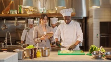 Profesyonel yetenekli Afro-Amerikan erkek şefin portresi sebze doğrayan ve tabletli güzel başarılı kadın yöneticiyle konuşan. Restoran mutfak konsepti.