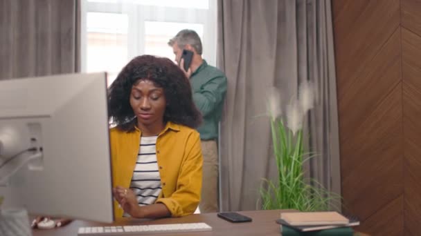 漂亮的非洲裔美国女性在电脑上工作 在键盘上打字 聊天和微笑 而漂亮的男性则站在后面 在手机上说话 看着窗户和女人 工作日概念 — 图库视频影像