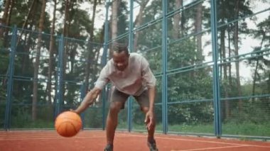 Açık hava sahasında basketbol oynayan olgun Afro-Amerikan erkek oyuncu. Yakışıklı yetişkin adam basketbol oynuyor ve zıplayan top antrenmanı yapıyor. Arka planda doğa, spor konsepti
