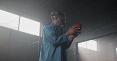 Çekici Afrikalı Amerikalı adam topu fırlatıyor ve karanlık odadaki kameraya bakıyor. Kasklı, spor giyimli yakışıklı bir erkek maça hazırlanıyor. Amerikan futbolu konsepti
