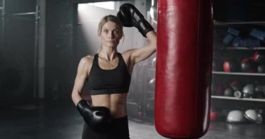 Spor merkezinde kum torbasının yanında duran yakışıklı beyaz kadın. Mükemmel kaslı vücudu olan güzel bir kadın kameraya bakıyor. Siyah takım elbiseli ve boks eldivenli güzel bir kız..