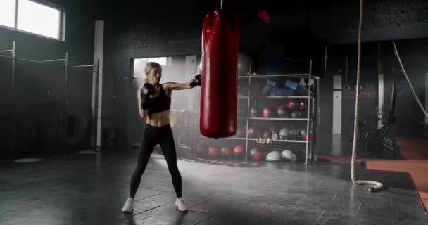漂亮的高加索女人在黑健身房练箱子 漂亮的女性看起来惊人与她的合适的身体 肌肉发达的女孩戴着手套打拳击袋 辛苦了 — 图库视频影像