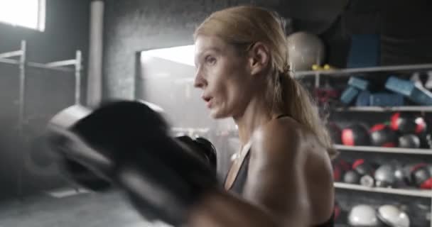 漂亮的高加索女人在体育馆里积极地打拳击袋 努力工作的女运动员 肌肉发达 准备挑战 背景中可见的运动物体 拳击运动的概念 — 图库视频影像