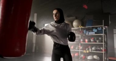 Beyaz kadın boks çantasına güçlü yumruklar atıyor. Koyu renk spor salonunda spor kıyafetleri ve eldivenleri olan güzel bir bayan antrenmanı. Boks ya da kickboks eğitimi. Spor Günü kavramı.
