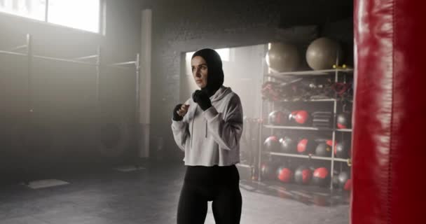 在白人拳击手女人的镜头中 站在拳打脚踢袋旁边看镜头 漂亮的女性 身穿运动衫 头戴帽衫 头戴手巾 看起来很有情趣 健身风格的概念 — 图库视频影像