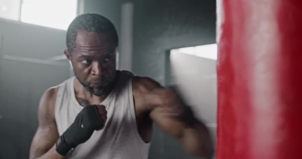 强壮的非裔美国人 带着拳击绷带在体育馆里锻炼 迷人的成年拳击手有力地冲撞悬挂式打斗袋 听着白色耳机中的音乐 — 图库视频影像
