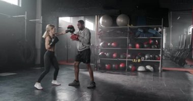 Sarı saçlı, spor takım elbiseli, boks eldivenli, spor salonundaki antrenörüyle antrenman yapan güzel bir kadın. İyi görünümlü, etnik çeşitlilikte insanlar birlikte çalışıyor. Boks sporu endüstrisi kavramı.