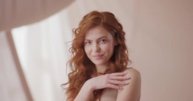 Kameraya bakan kıvırcık kızıl saçlı beyaz kadın portresi. Güzel bir bayan mutlu, göz kırpıyor, göz kırpıyor ve gülümsüyor, vücuda dokunuyor ve sağlıklı bir ten gösteriyor. Cazibe, cilt desteği, vücut bakımı.