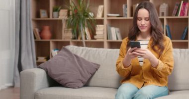 Güzel beyaz kadın rahat bir kütüphanede dinleniyor. Çekici bir kadın yumuşak koltukta oturuyor, cep telefonu kullanıyor ve gülümsüyor. Sevinçle mesaj yazmak, web iletişiminin tadını çıkarmak. Mobil aygıt kavramı.