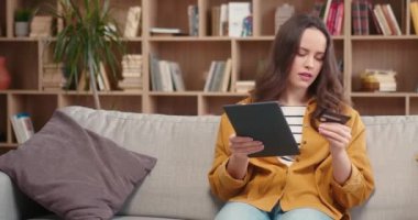 Tablet bilgisayarlı beyaz bir kadın uzaktan alışveriş yapıyor. Yumuşak koltukta oturan ve elinde kredi kartı olan genç bir kadın web sitesinden bir şey satın alıyor. İnternetten satın alma kavramı.