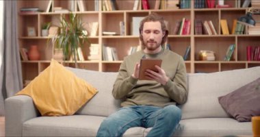 Yumuşak kanepede oturan ve elinde tablet tutan yakışıklı beyaz adam. Yakışıklı bir erkek kredi kartı kullanıyor ve internetten alışveriş yapıyor. Başarılı bir alışveriş yüzünden gülümsüyor. İnternet ödeme kavramı