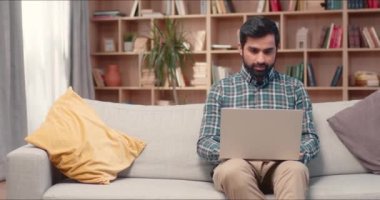 Modern ev kütüphanesinde dizüstü bilgisayar kullanan sakallı yakışıklı bir erkek. Kanepede oturan ve web sitelerinde veya sosyal medyada gezinen genç bir adam. Kameraya gülümsüyor, rahatlatıcı haftasonunun tadını çıkarıyor..