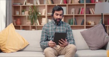 Ev kütüphanesinde çalışan sakallı yakışıklı bir adam. Genç yönetici yumuşak koltukta oturuyor, müşterilerle iş projesi hakkında video görüşmesi yapıyor. Tablet bilgisayar ve kulaklık kullanan meşgul erkek.