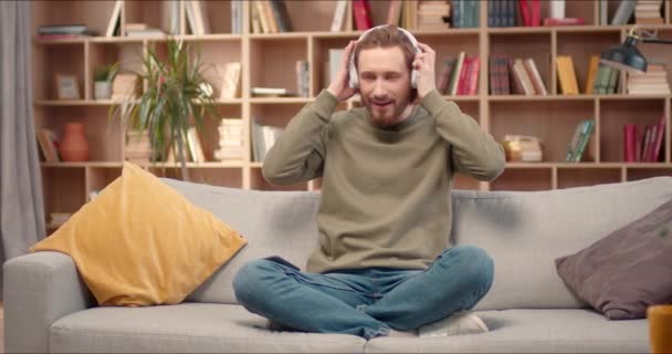 Vonzó Fehér Férfi Fülhallgatóval Könyvtárban Pihen Kényelmesen Lótuszban Pózol Kanapén — Stock videók