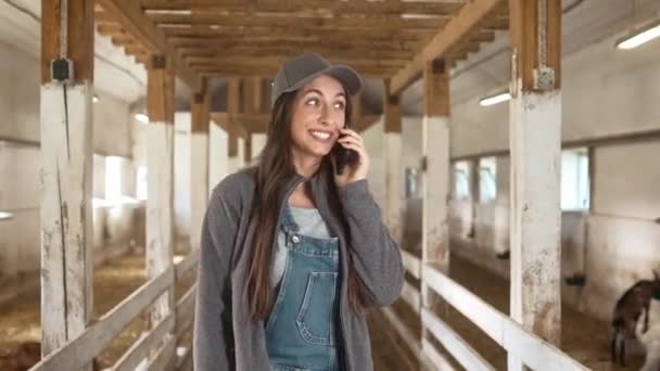 ヤギの養殖場を歩く愛らしい白人女性 喜んで電話で誰かと話す 魅力的な農家の笑顔と積極的な会話を持つ 女性の見える木製の小屋の後ろ — ストック動画