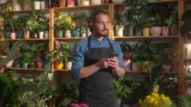 Önlüklü, beyaz tenli çekici bir işçi elinde telefonla birilerine mesaj yazıyor. Arka planda yeşil bitkilerle dolu ahşap raflar var. Çiçekçide çalışıyorum. Teknoloji kullanılıyor.