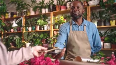 Müşteri çiçek buketini kredi kartıyla öderken önlük taşıyan mutlu Afrikalı Amerikalı satıcı. Adamın arkasında bitkiler ve organik otlarla dolu raflar var. Satın alma. Ödeme.