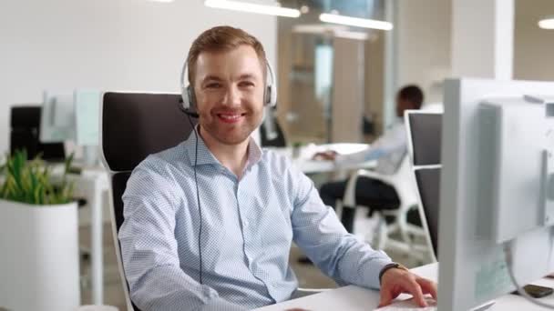 放大到快乐的白人男性操作员坐在办公室的计算机前 积极的员工 带着耳机支持客户 并在镜头前微笑 呼叫中心服务的概念 — 图库视频影像