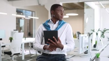 Ofisteki bilgisayar dolu masada oturan çekici Afrikalı Amerikalı adam. Seçkin yönetici, tablet ve müşteriyle uzaktan mesajlaşma kullanıyor. Ticaret şirketinde çalışmaya odaklanmış..