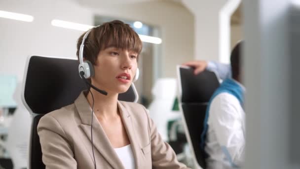 忙碌的白人操作员女人带着耳机和客户谈论股票市场 舒适地坐在现代办公室的计算机前 两名多族裔男子进行交流 — 图库视频影像