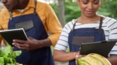 İki Afrikalı Amerikalı çiftçinin portresi organik hasatları sayarken yiyeceklerin arkasında duruyor. Not defterinde satılan ürün sayısını yazmakla meşgulken tablet aygıtı kullanan güzel kadın.