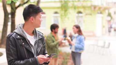 Kasabanın caddesinde cep telefonuyla yazan deri ceketli mutlu Asyalı adamın portresini yakından çek. Genç adam akıllı telefondan internette geziniyor. Arka plandaki arkadaşlar, şehir tarzı konsepti