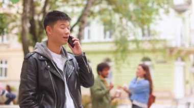 Asyalı neşeli genç adam akıllı telefonla konuşuyor ve dışarıda dururken gülümsüyor. Caddede cep telefonuyla konuşan yakışıklı adamın portresi. Olumlu duygular, iletişim
