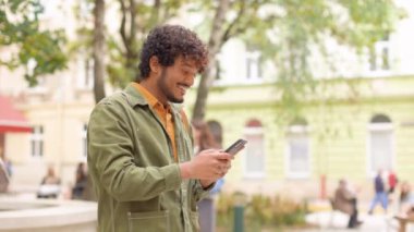 Açık havada akıllı telefondan mesajlaşan mutlu genç erkek portresi. Yakışıklı kıvırcık adam sokakta cep telefonuyla yazı yazıyor. Kentsel yaşam tarzı, olumlu duygular, boş zaman kavramı