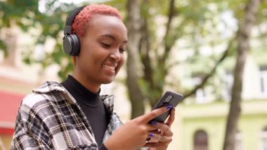 Mutlu gülümseyen genç Afrikalı Amerikalı kadının cep telefonuyla neşeli bir şekilde mesajlaşmasını kapatın. Kulaklıkla müzik dinleyen, akıllı telefondan internette sörf yapan mutlu, güzel bir kadın.