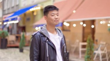 Kamera şehir merkezinde kameraya gülümseyen yakışıklı Asyalı adama yaklaşıyor. Şehir merkezinde mutlu yüzlü bir erkek turist var. Boş zaman, turizm, şehir tarzı, yakın çekim.