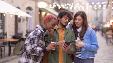 Dekorasyonlu caddede durup cep telefonuyla ekranlara bakan neşeli, çoklu etnik kökenli genç arkadaşların portresi. Cep telefonu ve turizm kullanan mutlu erkek ve kadın turistler