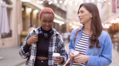 Kentin süslü sokaklarında ellerinde kahveyle yürüyen ve konuşan çok ırklı genç güzel bayan arkadaşlarını yakından takip edin. Konuşan ve gülen kadınlar. Kahve zamanı, iletişim konsepti.