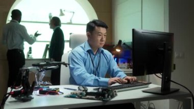 Laboratuvarda bilgisayar üzerinde çalışan Asyalı geliştirici. Teknik cihazlar masanın üzerinde. İş arkadaşları birbirleriyle iletişim kurarken klavyede bir şeyler yazmakla meşgul..