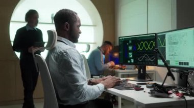 Laboratuvarda yenilik projesi üzerinde çalışan Afrikalı Amerikalı mühendisin yan görünüşü. Mesgul bir adam iskemlede oturup kod veya formül yazmakla meşgul. Görünürde hologramlar ve devreler var. İşbirliği.