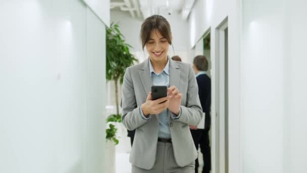 同僚が立っている白い廊下を歩く美しい白人女性マネージャー スマートフォンや誰かとのメッセージを使っている若い女性 陽気な笑顔 気持ちいい — ストック動画
