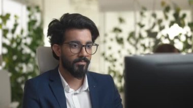Bilgisayarın önünde oturan gözlüklü ve takım elbiseli genç sakallı yönetici. Özenle PC monitörüne bakıyor ve gülümsüyor. Modern ofiste çalışan çekici bir adam. İş konsepti.