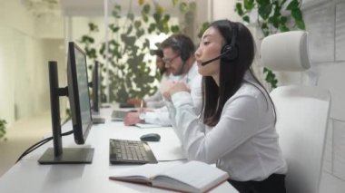 Modern ofisteki müşterilerle iletişim kuran kulaklıklı çekici Asyalı bayan yönetici. Müşterileri arayan meşgul operatörlerin yanında. Bankacılık ticareti. İş şirketi.