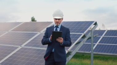Dışarıda güneş paneli arka planında profesyonel müdürün portresi var. Takım elbiseli ve kasklı bir erkek işadamı görev yapmak için tablet kullanıyor. Yeşil elektrik kavramı.
