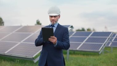 Kasklı ve takım elbiseli endüstriyel yönetici tablet kullanıyor ve güneş panelleri üretiminde teftiş yapıyor. Profesyonel sakallı iş adamı dışarıda çalışıyor. Yeşil elektrik kavramı.