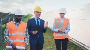 Dışarıdaki güneş paneli arka planında üç çok kültürlü işçi yürüyor ve işbirliği yapıyor. Yansıtıcı yelek ve kask giyen çalışanlar iş yerinde iletişim kuruyor. Yeşil elektrik kavramı.