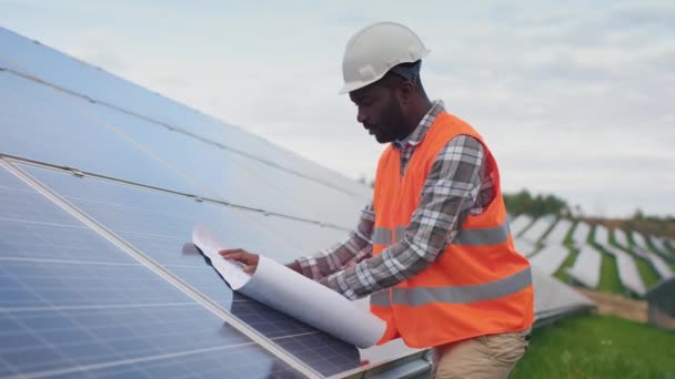 一个从事太阳能电池板生产的非裔美国人的肖像 穿着防护背心和硬礼帽的工人在外面看并查看图纸 绿色电力的概念 — 图库视频影像