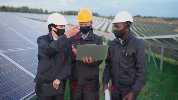 戴面具的工程师站在太阳能电池板的后面 成年男子穿着防护服 头戴硬帽 使用笔记本电脑 并与同事交流 非裔美国人持有蓝图 — 图库视频影像