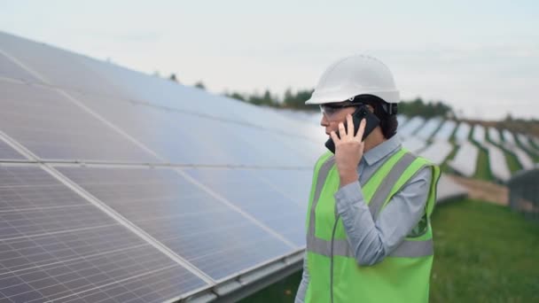 穿着反光背心 头盔和谷歌眼镜的女员工正在外面检查太阳能电池板的生产情况 女人与同事交流 并通过电话交谈 绿色电力的概念 — 图库视频影像