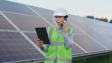 Spesiyal üniformalı kadın işçinin portresi ve arka planda güneş panelleri olan sert bir şapka. Ekoloji mühendisi telefonla ve dışarıda tablet kullanımında çalışıyor. Yeşil elektrik kavramı.