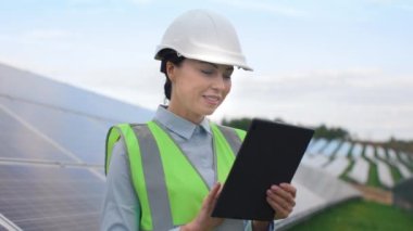 Spesiyal üniformalı bir iş kadını portresi ve arka planda güneş panelleri olan bir şapka. Kadın işçiler işlerinden memnun. Kadın dışarıda tablet kullanıyor. Yeşil elektrik kavramı.
