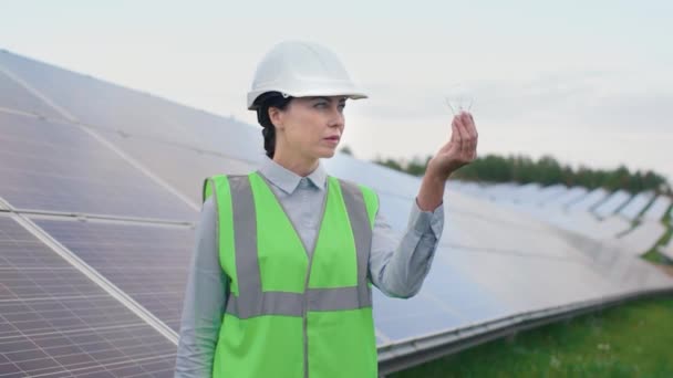 緑の電気の概念 ソーラーパネルの太陽の背景に立つ女性エンジニア ハード帽子と反射ベストを身に着けている女性は電球を保持しており 笑顔でカメラを見ています — ストック動画