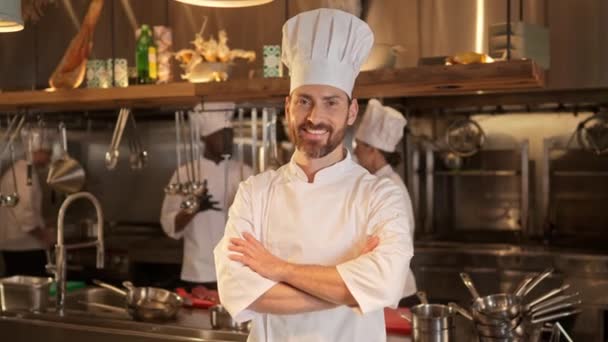 笑顔の男性シェフは レストランのキッチンでの作業工程を背景に腕を組んで立っています 一緒に働く特別な制服の多文化料理人のチーム — ストック動画