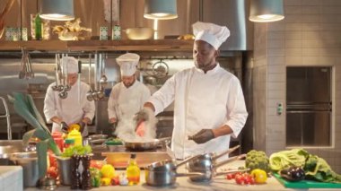Afro-Amerikalı aşçı, ticari restoranın mutfağında lezzetli yemekler hazırlıyor. Çok kültürlü bir şef derin tavada baharatlı et yakar. Profesyonel dumanlı yemek pişirme.